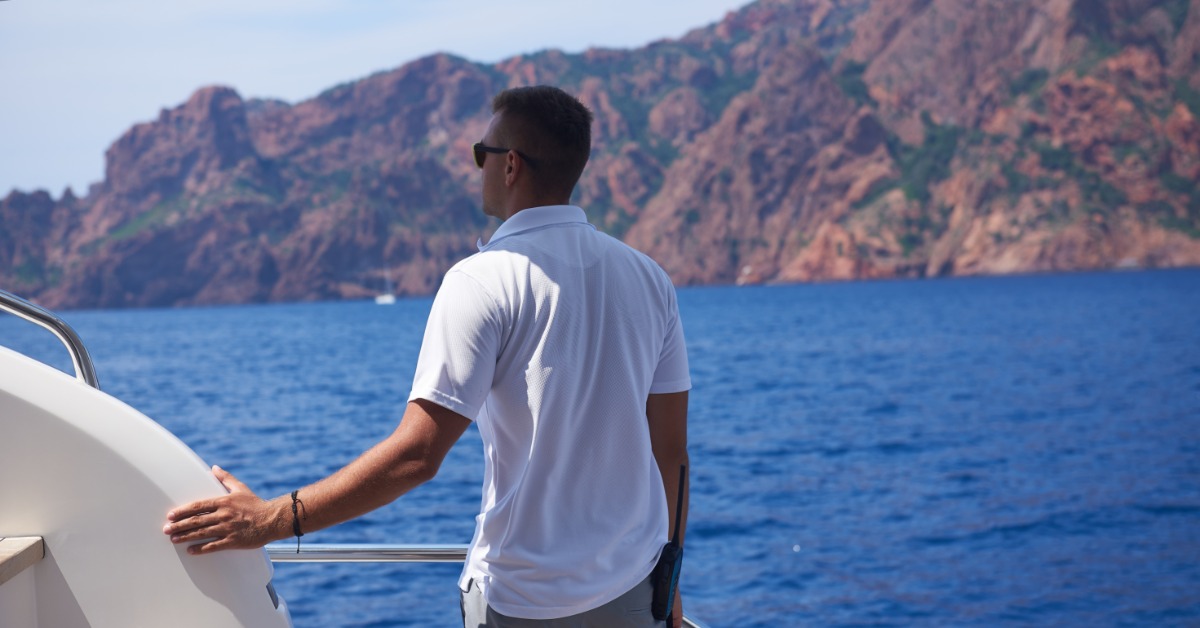 A man wearing a white polo and grey shorts, standing in the back of a yacht overlooking the water and a mountain.